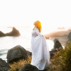 Eine Frau in einem weißen Kleid genießt die frische Brise an einer wilden Küste bei Sonnenuntergang. Ein Moment purer Freiheit, Naturverbundenheit und magischer Kraft – die Essenz von BREEZE einer ESSENCE-ORIGINALS Duftmischung.